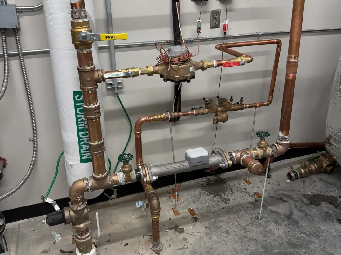 Copper water piping with a water meter, a yellow-handled shutoff valve and a backflow preventer on a mechanical room wall